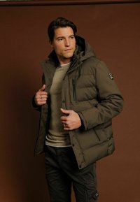 Olijfgroene pufferjas met een capuchon, ritszakken en logo patch. Draag het over een beige trui en donkere broek. Glad textuur.