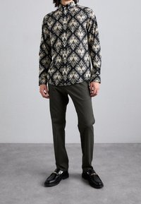 Camicia nera a maniche lunghe con un motivo barocco dorato, abbinata a pantaloni verde oliva e mocassini neri con dettagli in oro.