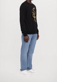 Man i svart sweatshirt med guldig ormsgrafik, ljusblå jeans och vita sneakers, stående mot en enfärgad vit bakgrund.