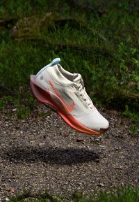 Chaussure de course Nike en crème, avec des accents orange et rose. Caractéristiques d'une tige respirante, d'une semelle texturée et d'un design épuré.