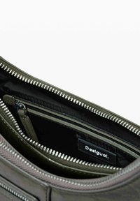 Umhängetasche mit einem strukturierten, dunkelgrünen Äußeren, Nylon-Innenfutter und mehreren Fächern mit metallischen Reißverschlussakzenten. Label sichtbar.