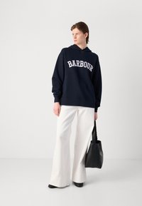 Moletom azul marinho com o logótipo "BARBOUR" em branco, combinado com calças de perna larga brancas e uma mala preta. Tecido suave, design casual.