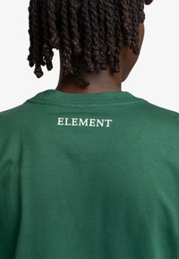 Grøn bomulds T-shirt med rund hals og korte ærmer. På bagsiden er "ELEMENT" trykt i hvid. Glat tekstur med minimale designdetaljer.