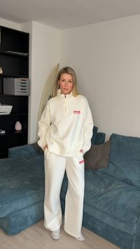 Γυναίκα με λευκή φούτερ Rip Curl και παντελόνι στέκεται με τα χέρια στις τσέπες σε μια σαλονοτραπεζαρία δίπλα σε μια μπλε καναπέ και μια μαύρη μονάδα ραφιών.