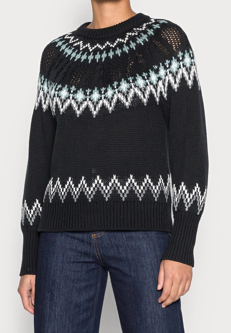 Pull en maille noir avec des motifs en zigzag et en losanges blancs et bleu clair sur la poitrine, les manches et l’ourlet, porté avec un jean bleu.