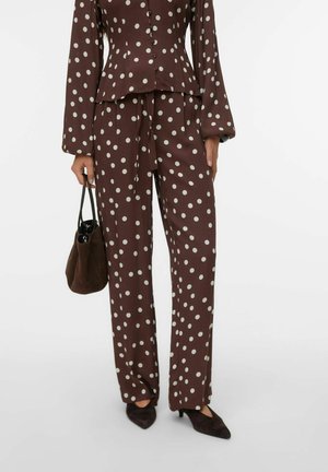 Bruin set van een blouse met lange mouwen en broek met witte polkadots, gecombineerd met zwarte puntige hakken en een donkerbruine handtas in de linkerhand.