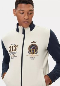 Giacca in pile color crema e blu con collo alto, zip centrale e dettagli ricamati tra cui un emblema e numeri sulla parte anteriore.