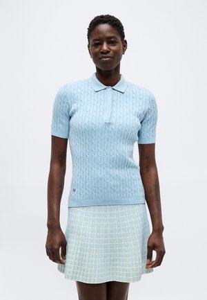 Frau trägt ein hellblaues kurzärmeliges kabelgestricktes Poloshirt und einen gelb-hellblau karierten Rock, steht vor weißem Hintergrund.