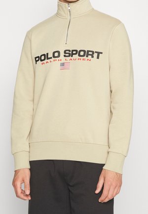 Beżowa bluza z suwakiem do połowy klatki piersiowej z napisami „POLO SPORT” i „RALPH LAUREN” nad małą amerykańską flagą, noszona z czarnymi spodniami.