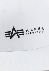 Căciulă albă cu un logo negru, cu dungi și textul "ALPHA INDUSTRIES". Materialul pare neted, cu o coroană structurată și o vizieră curbată.