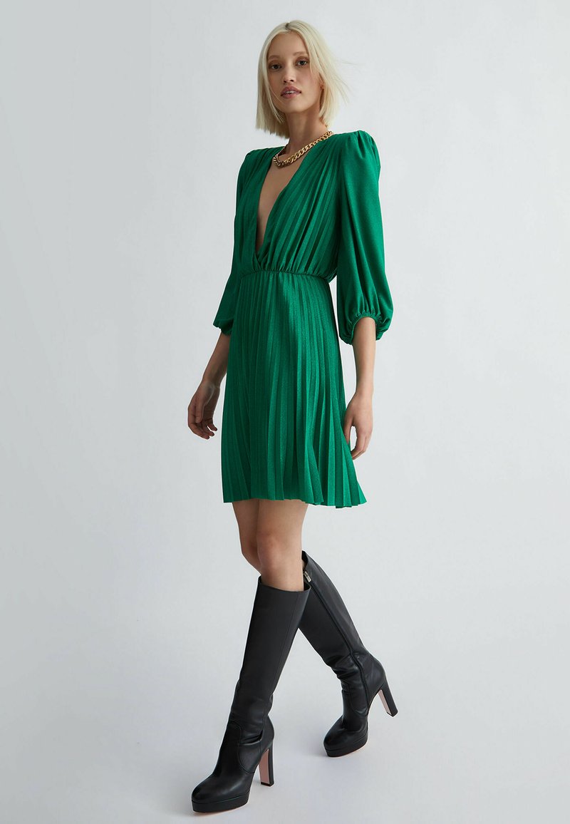 Grünes plissiertes Kleid mit tiefem V-Ausschnitt und Puffärmeln, kombiniert mit hohen schwarzen Lederstiefeln mit Absätzen. Das Kleid hat eine figurbetonte Taille.