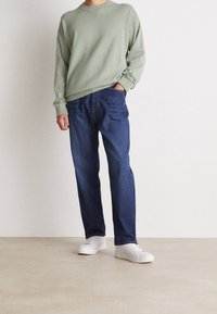 Lichtgroene trui met geribde halslijn, gecombineerd met donkerblauwe straight-leg spijkerbroek. Het model draagt witte sneakers, wat een casual uitstraling benadrukt.