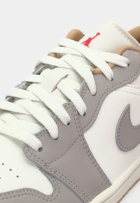 Gros plan d'une basket blanche et gris clair avec des lacets blancs et un logo rouge sur la languette, montrant des coutures détaillées et des perforations.