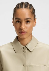 Junge Frau mit geflochtenem Haar, die ein beige-knöpfbares Ledershirt trägt, schaut mit neutralem Ausdruck auf einem einfarbigen Hintergrund nach vorne.