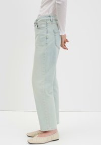 Hellblaue, hoch taillierte Jeans mit geradem Bein, Fronttaschen und minimalen Nähdetails, kombiniert mit beigen Schlupfschuhen.