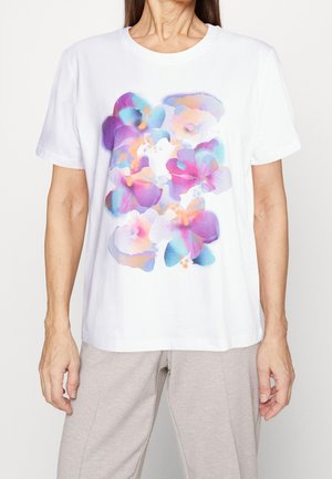 Femme portant une chemise blanche à manches courtes avec un imprimé floral abstrait coloré en nuances de violet, bleu et rose.