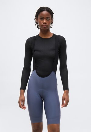 Γυναίκα με μαύρη μακρυμάνικη βασική μπλούζα ποδηλασίας και μπλε ποδηλατικά σορτς με ψηλή μέση, μπροστά σε απλό φόντο.