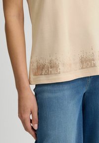 Beige T-shirt met korte mouwen en een decoratief, geperforeerd ontwerp aan de onderkant. Het materiaal is zacht met een gladde textuur en wordt gecombineerd met denim.