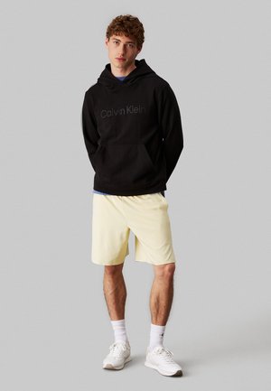 Felpa nera in cotone con tasca frontale e logo, abbinata a pantaloncini beige. Il modello è in piedi con le braccia incrociate, indossa scarpe da ginnastica bianche e calzini.