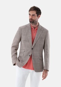 Hellbrauner karierter Blazer mit Revers, zwei vorderen Taschen, kombiniert mit einem korallefarbenen Poloshirt. Weiße Hose und dunkle Knöpfe vervollständigen das Outfit.