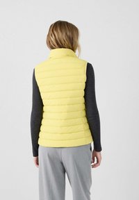 Chaleco acolchado amarillo con diseño de acolchado horizontal, cuello alto y forma ajustada, usado sobre una camiseta de manga larga gris oscuro y pantalones gris claro.
