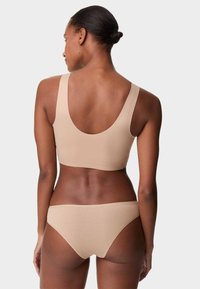 Mujer de piel oscura y cabello recogido en un moño, vestida con un sujetador beige sin costuras y ropa interior a juego, de pie de espaldas contra un fondo liso.