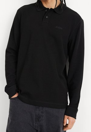 Man wearing black long-sleeve polo shirt with "BOSS" logo on chest and dark gray jeans, hand in pocket, against plain background.