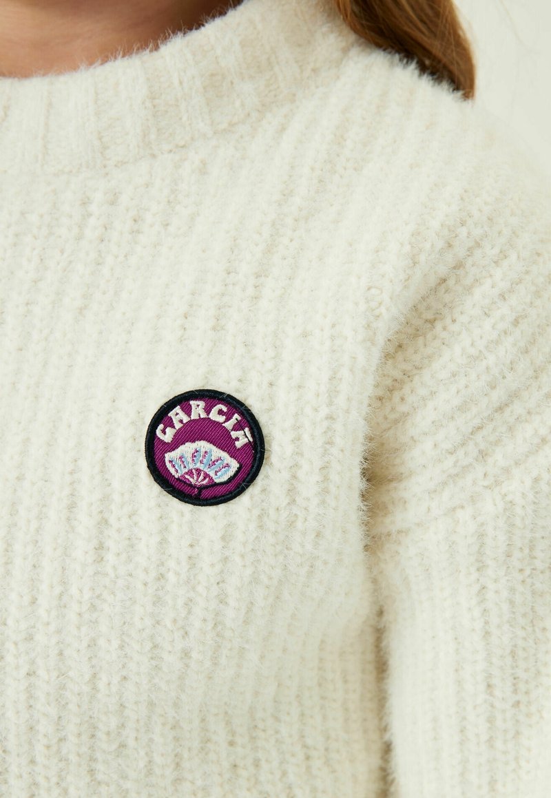 Gros plan sur un pull côtelé de couleur crème avec un écusson rond violet montrant un éventail et le mot « GARCIA » sur la poitrine.