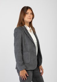 Giovane donna con lunghi capelli castani, che indossa un completo in velluto grigio e una camicia bianca, in piedi leggermente girata verso uno sfondo chiaro e semplice.