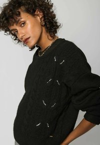 Pull en tricot noir à mailles larges avec une coupe décontractée, présentant des motifs texturés et des accents métalliques, porté sur une chemise à col.