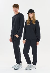 Donkerblauwe sweatshirt en joggers, gemaakt van zachte stof. De joggers hebben zakken, en beiden dragen lichtgekleurde sportschoenen. Een zwarte pet wordt door één persoon gedragen.