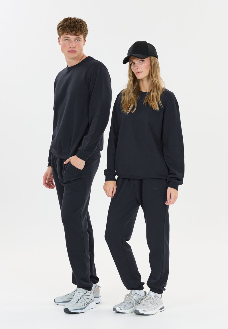 Donkerblauwe sweatshirt en joggers, gemaakt van zachte stof. De joggers hebben zakken, en beiden dragen lichtgekleurde sportschoenen. Een zwarte pet wordt door één persoon gedragen.