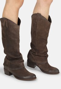 Stivali alti in suede marrone con altezze morbide, punta tonda e tacco basso impilato. Presentano una finitura liscia e dettagli di cucitura minimi.