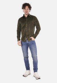 Camicia verde oliva a maniche lunghe con design a bottoni, realizzata in tessuto morbido. Indossata con jeans blu e sneaker bianche.