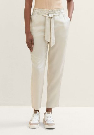 Pantaloni beige chiaro con cintura in vita, caratterizzati da una texture liscia e tasche laterali. Abbinati a sneakers bianche con accenti beige.
