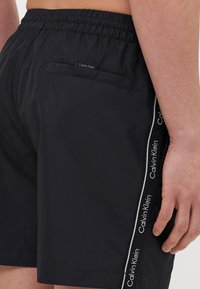 Fekete úszónadrág gumis derékkal és hátsó zsebbel. Az oldalsó csíkokat fehér "Calvin Klein" logó díszíti, kontrasztos háttéren.