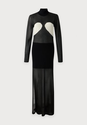 Femme aux cheveux blonds raides porte une robe noire transparente avec des formes de cœur blanches sur la poitrine et des talons noirs, debout sur un fond uni.