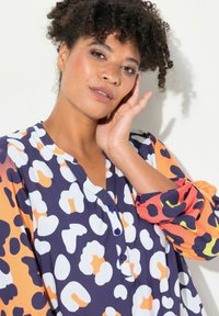Blusa colorata con base blu navy e motivi astratti bianchi, arancioni e verdi. Presenta un'apertura con bottoni e maniche arricciate.