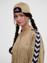 Mlada ženska z dolgimi pletenicami nosi bež vzorčasto jakno z zadrgo in ujemajočo se obratno baseball kapo z mornarsko modrimi cik cak črtami na rokavih.