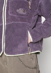 Giacca in pile viola con chiusura a zip, caratterizzata da una texture morbida, dettagli in crema e un logo sulla tasca del petto. Tasche laterali con zip.