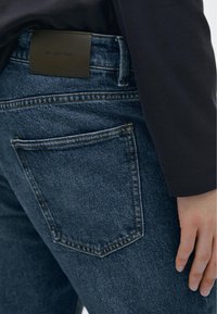 Modré džínové kalhoty mají zadní kapsu s viditelným prošíváním a obdélníkovou hnědou koženou cedulku v pase s logem "Massimo Dutti".