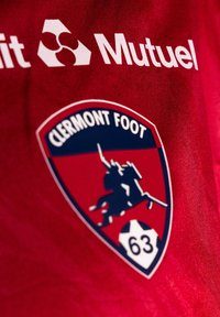 Röd sporttröja med Clermont Foot-logotypen. Logotypen har en sköldform med en silhuett av en ryttare och en fotboll, detaljerad i blått och vitt.