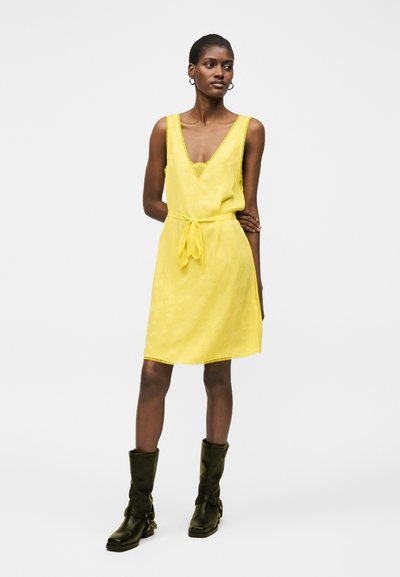 Γυναίκα στέκεται με σταυρωμένα τα χέρια πίσω από την πλάτη, φορώντας ένα κίτρινο φόρεμα χωρίς μανίκια με ζώνη στη μέση και μαύρα μπότες μέχρι τη μέση της γάμπας σε λευκό φόντο.