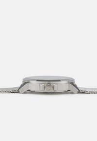 Reloj de metal plateado con una esfera negra redonda, botón de presión texturizado y correa de malla. Presenta un acabado pulido y detalles claros en la caja.