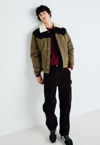 Veste matelassée verte et marron avec un col en fausse fourrure, portée sur un haut bordeaux, associée à un pantalon en velours côtelé noir et des baskets noires.