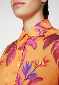 Cămașă portocalie cu modele florale mari în nuanțe de roz și mov, cu guler butonat și un mic detaliu auriu în partea stângă.