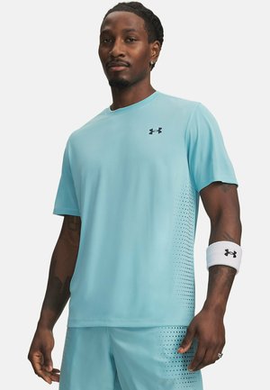 Hombre con camiseta deportiva y pantalones cortos Under Armour azul claro con lados perforados, banda blanca en la muñeca, de pie frente a un fondo liso.