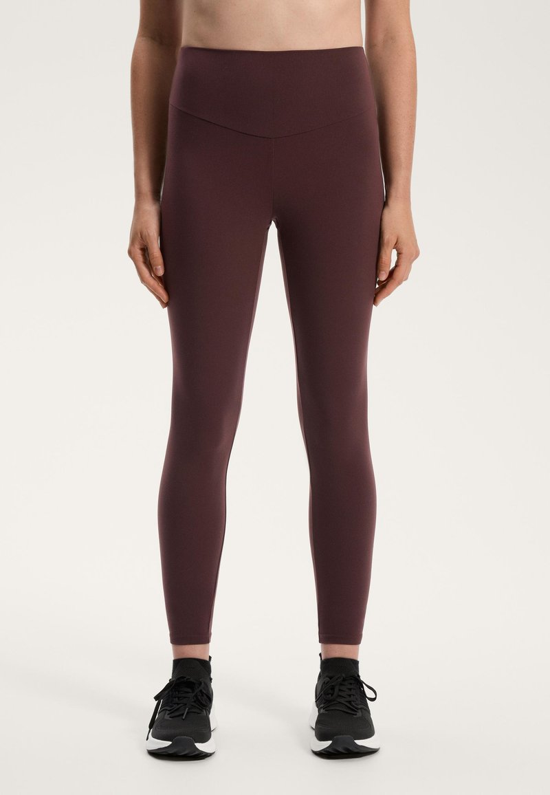 Bordo visokoslojni leggingi iz raztegljive tkanine, ki imajo gladko teksturo in prilegajočo se zasnovo, v kombinaciji s črnimi športnimi čevlji.