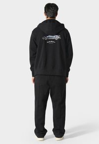 Felpa nera con cappuccio caratterizzata da una grafica di un'auto sul retro, con testo bianco sottostante. Abbinata a pantaloni neri e scarpe chiare.