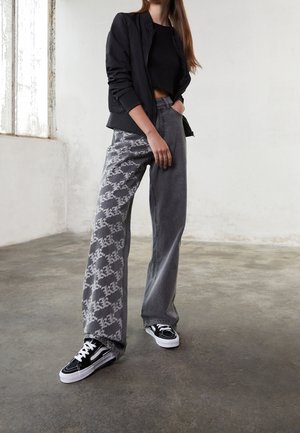 Woman in black cropped top, black jacket, wide-leg gray jeans with white pattern on one leg, and black and white sneakers standing indoors.